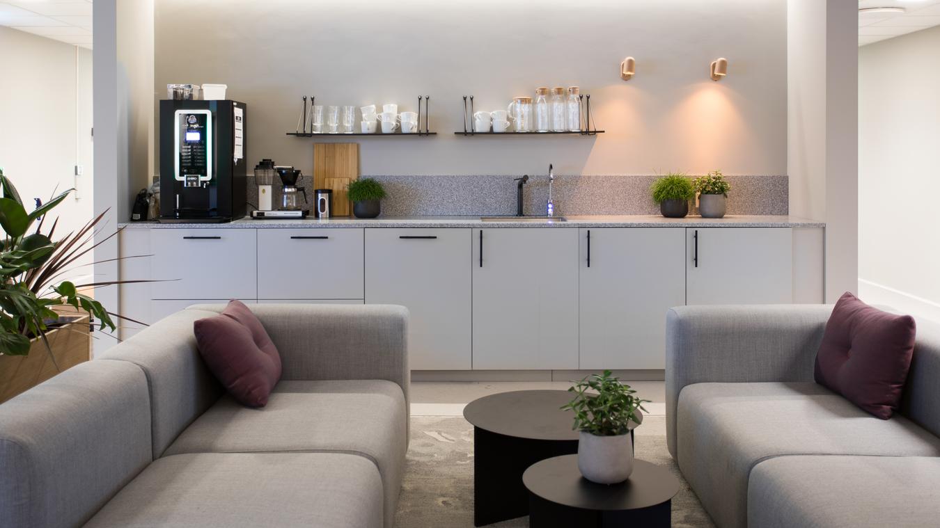 Kjøkken i kontorlandskap med grå detaljer og sittegruppe med sofa og bord. Foto