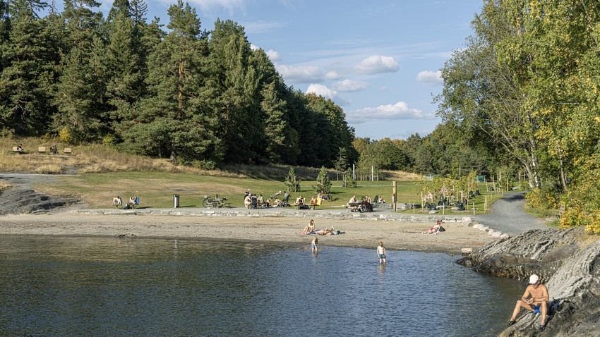 Badebukt på en varm sommerdag. Strand med grønn skog bak. Badende og solende folk. Foto