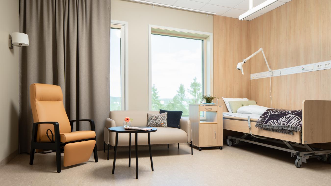 Soverom på sykehjem i lune oransje og grå farger. Sittegruppe og seng. Foto